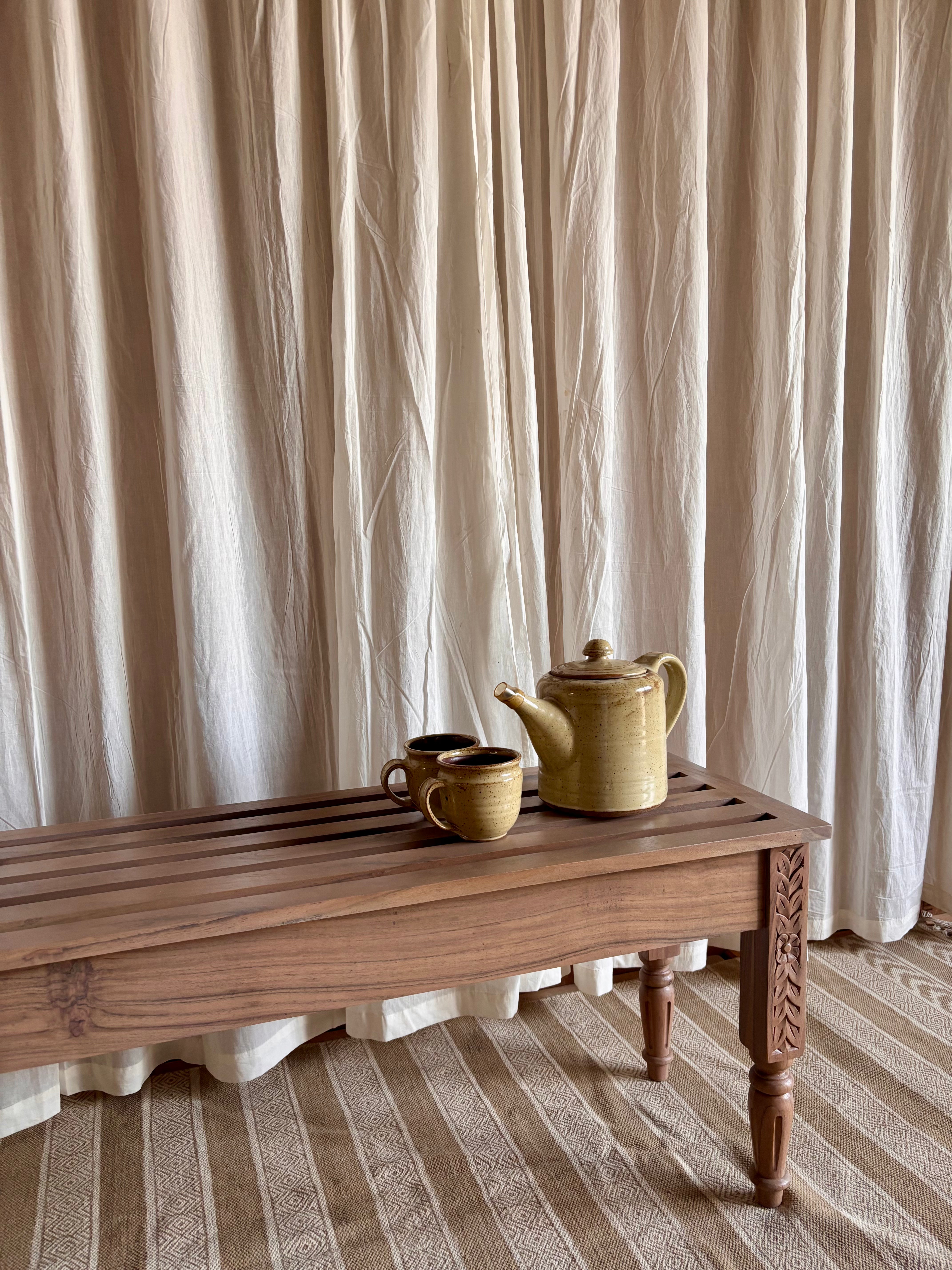 Teapot and cups on a wooden bench with beige curtains in the background